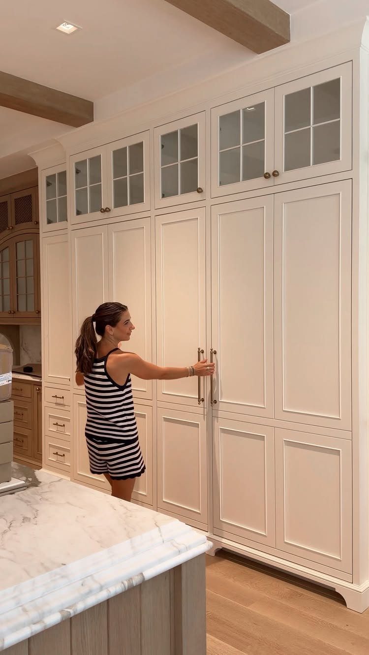 Woman opening a large white built-in hutch with glass upper cabinets and brass hardware.
