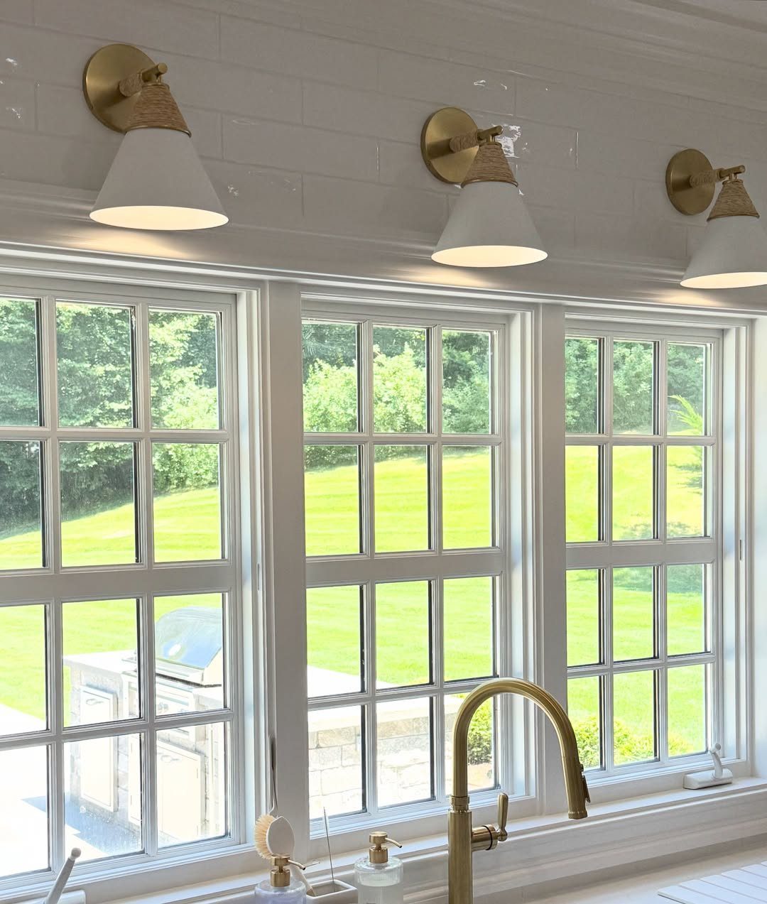 Three white conical wall sconces with brass mounts installed above white kitchen windows.