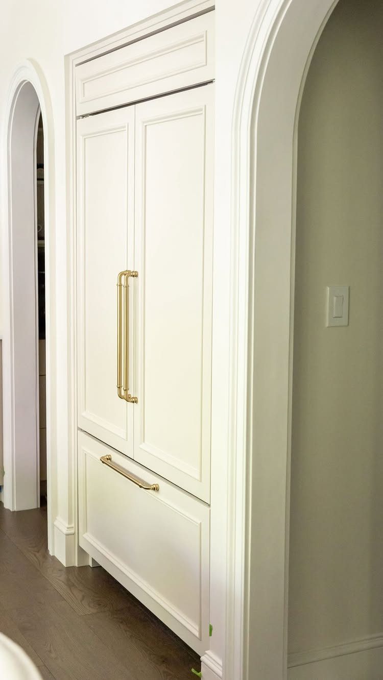 White integrated French-door refrigerator with gold handles built into a white cabinet.
