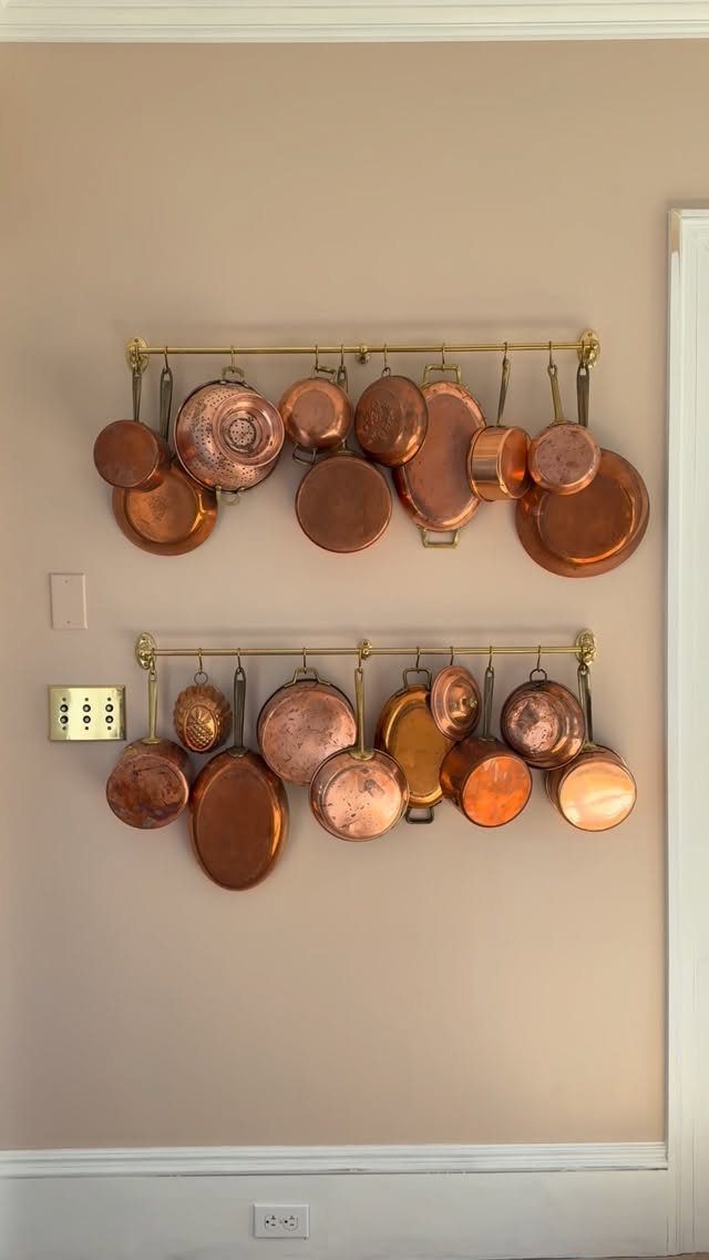 Copper pots and pans hanging from two brass rails on a beige wall.