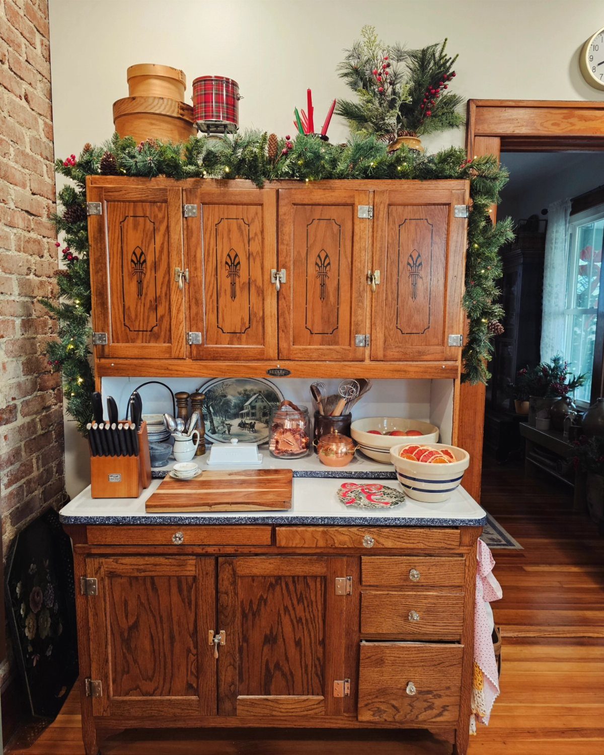 Vintage wooden Hoosier cabinet decorated with festive evergreen garlands and warm white lights.