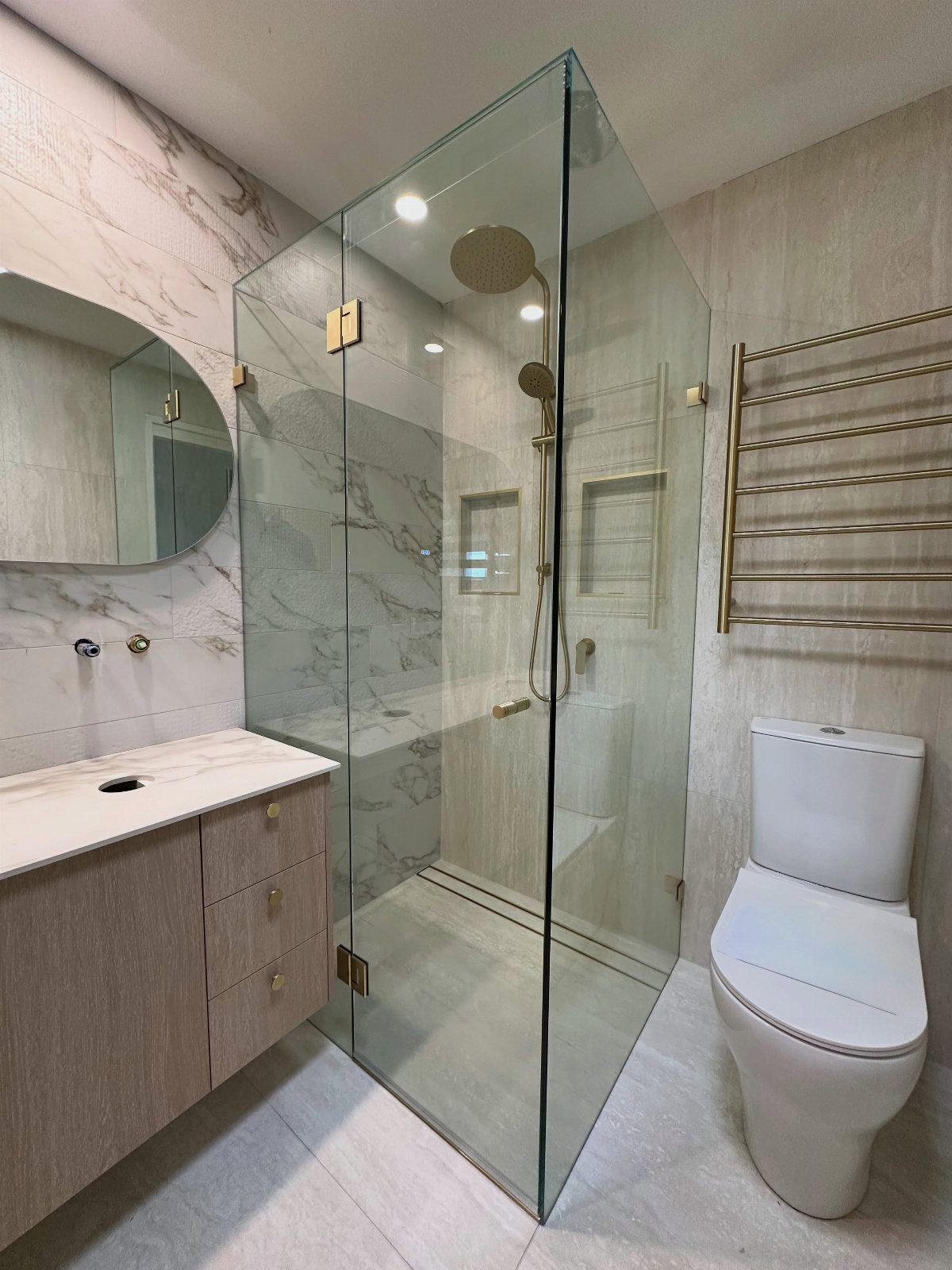 Frameless glass shower panels in a modern bathroom with marble walls and gold fixtures.