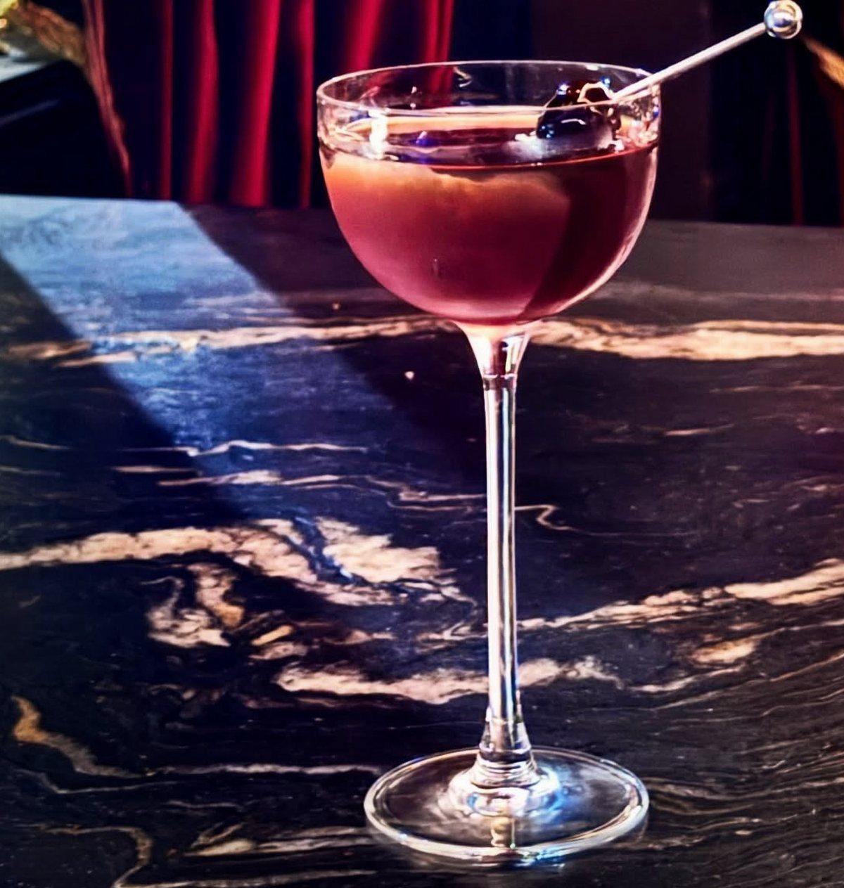 Red cocktail in a coupe glass on a polished black marble bar top