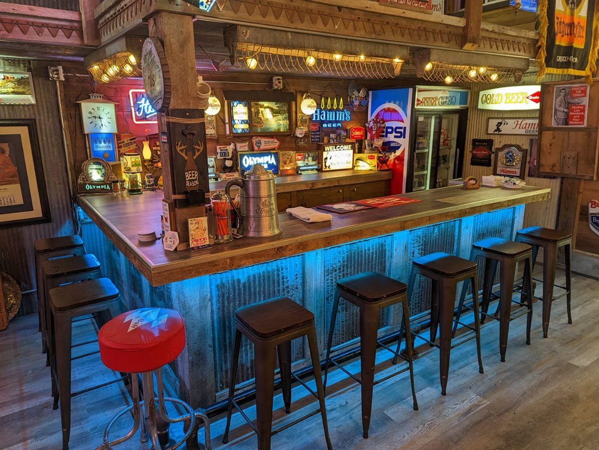 Rustic indoor bar with blue under-lighting, wooden countertop, and various vintage beer signs.