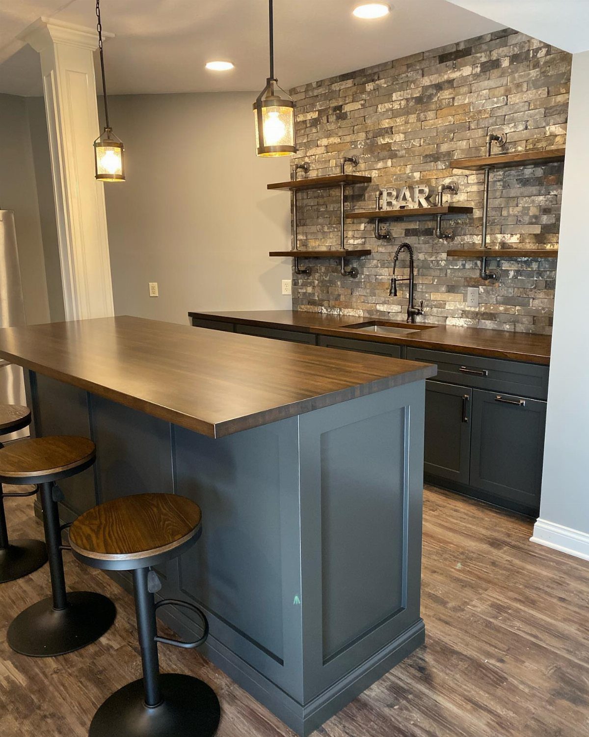Dark grey home bar with wooden countertops, stone backsplash, and floating shelves.