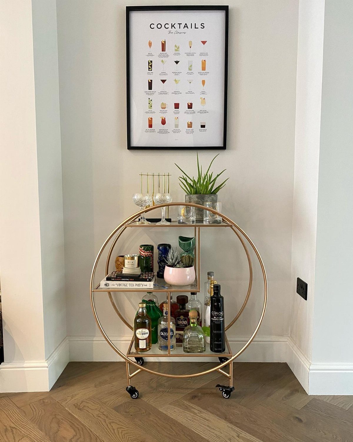 Gold circular bar cart with liquor bottles and glassware against a white wall.