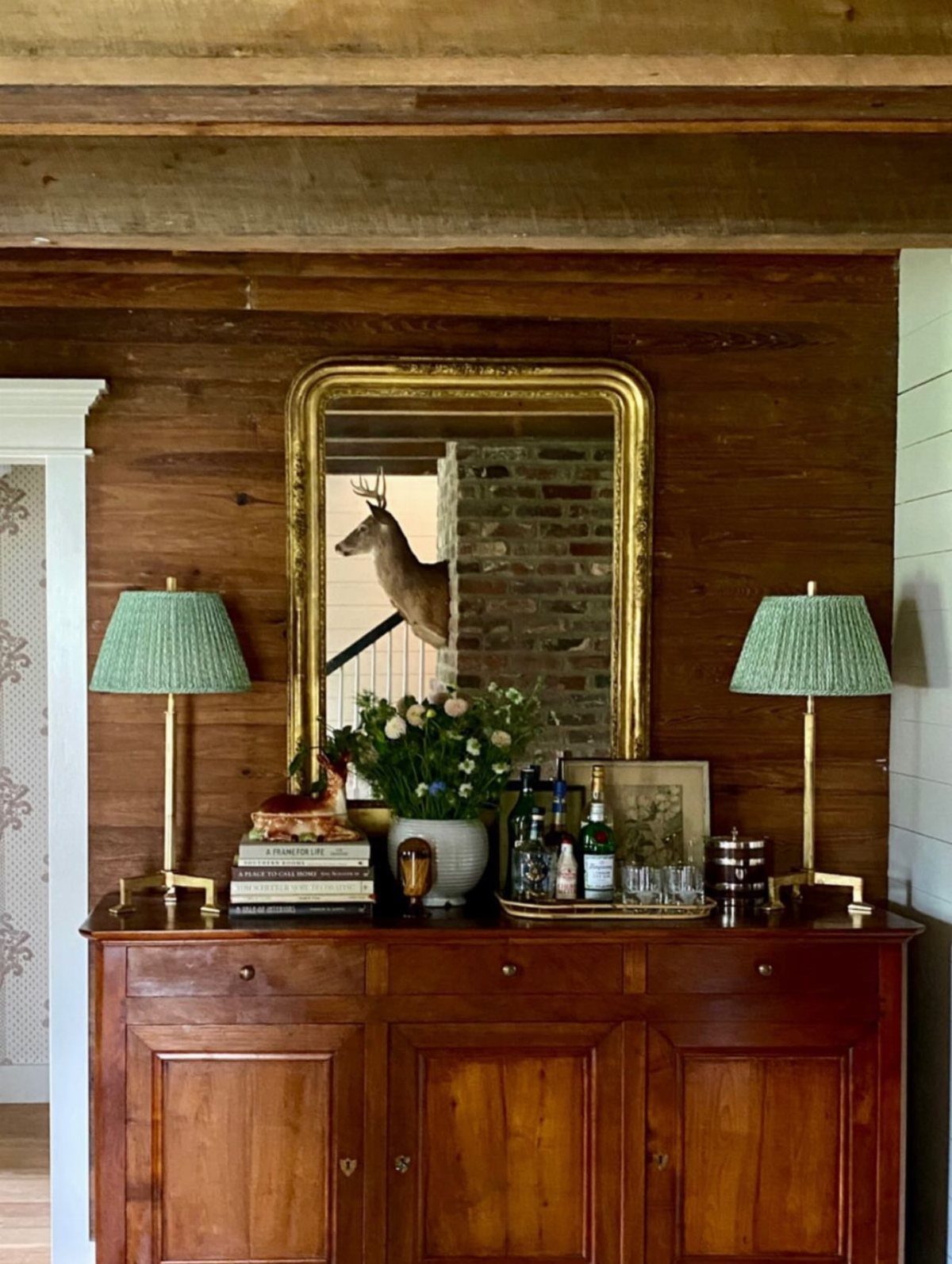 Wooden sideboard bar with green lamps, gold mirror, and liquor bottles on tray.