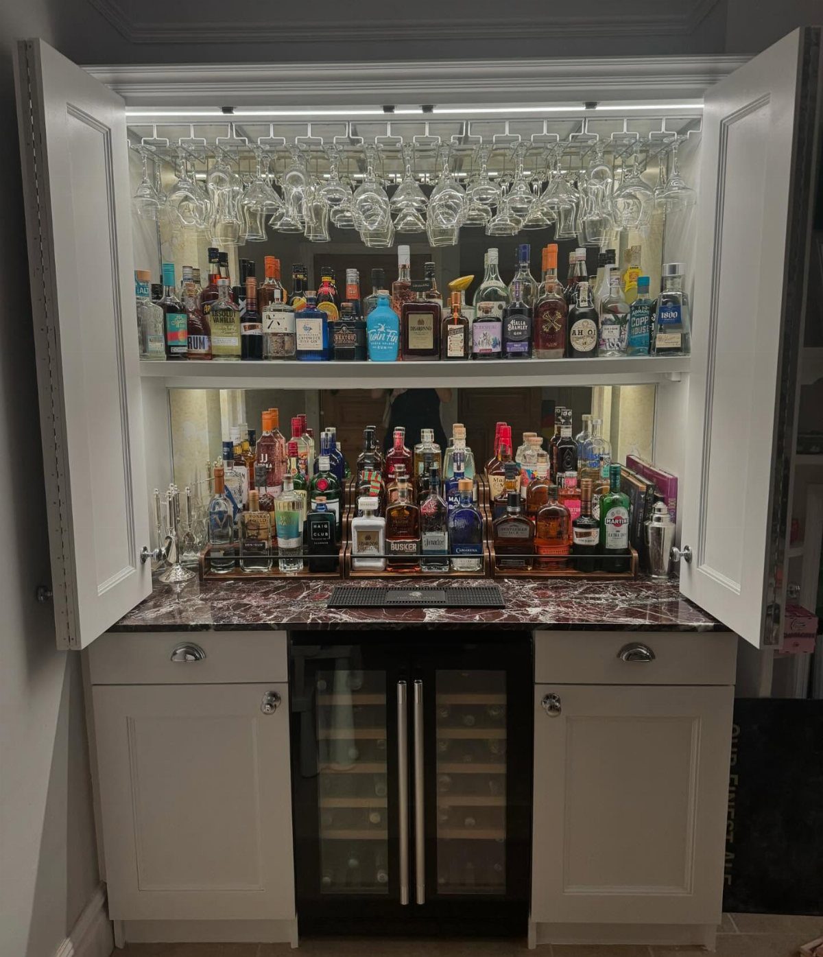White built-in home bar with marble countertop, wine cooler, and stocked liquor shelves.