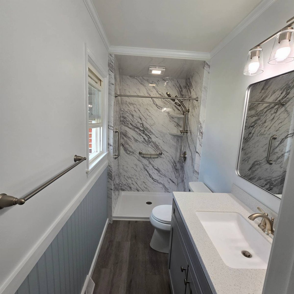 Small bathroom with marble walk-in shower, grey vanity, and light blue wainscoting walls.