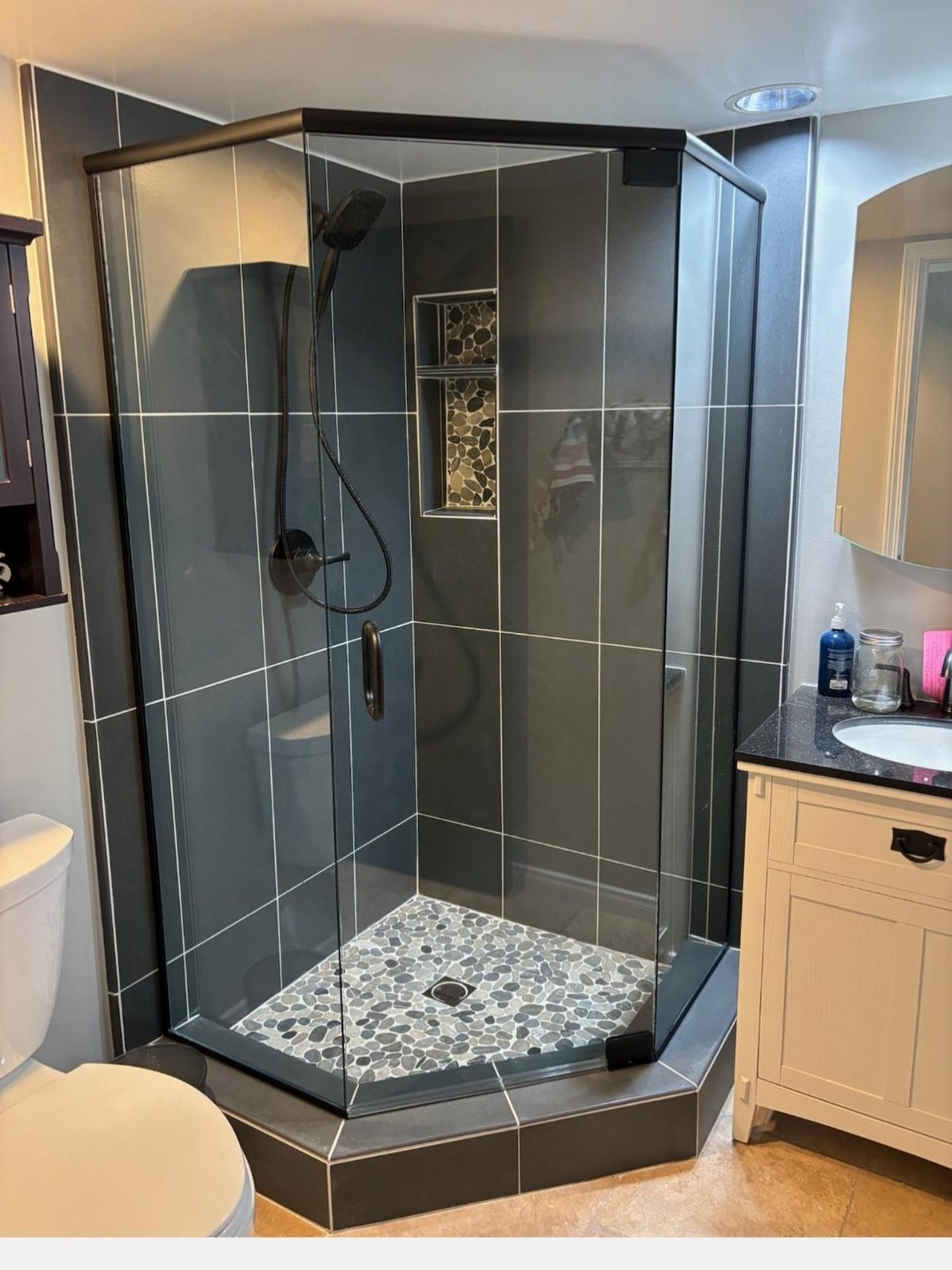 Corner shower with black glass frame, grey tiles, and pebble floor in a bathroom.