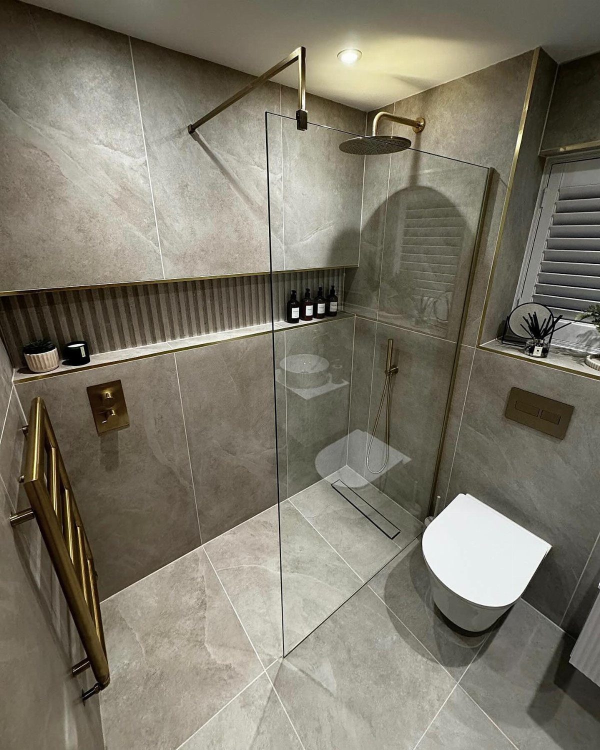 Curbless walk-in shower with glass panels and gold fixtures in a grey marble bathroom.