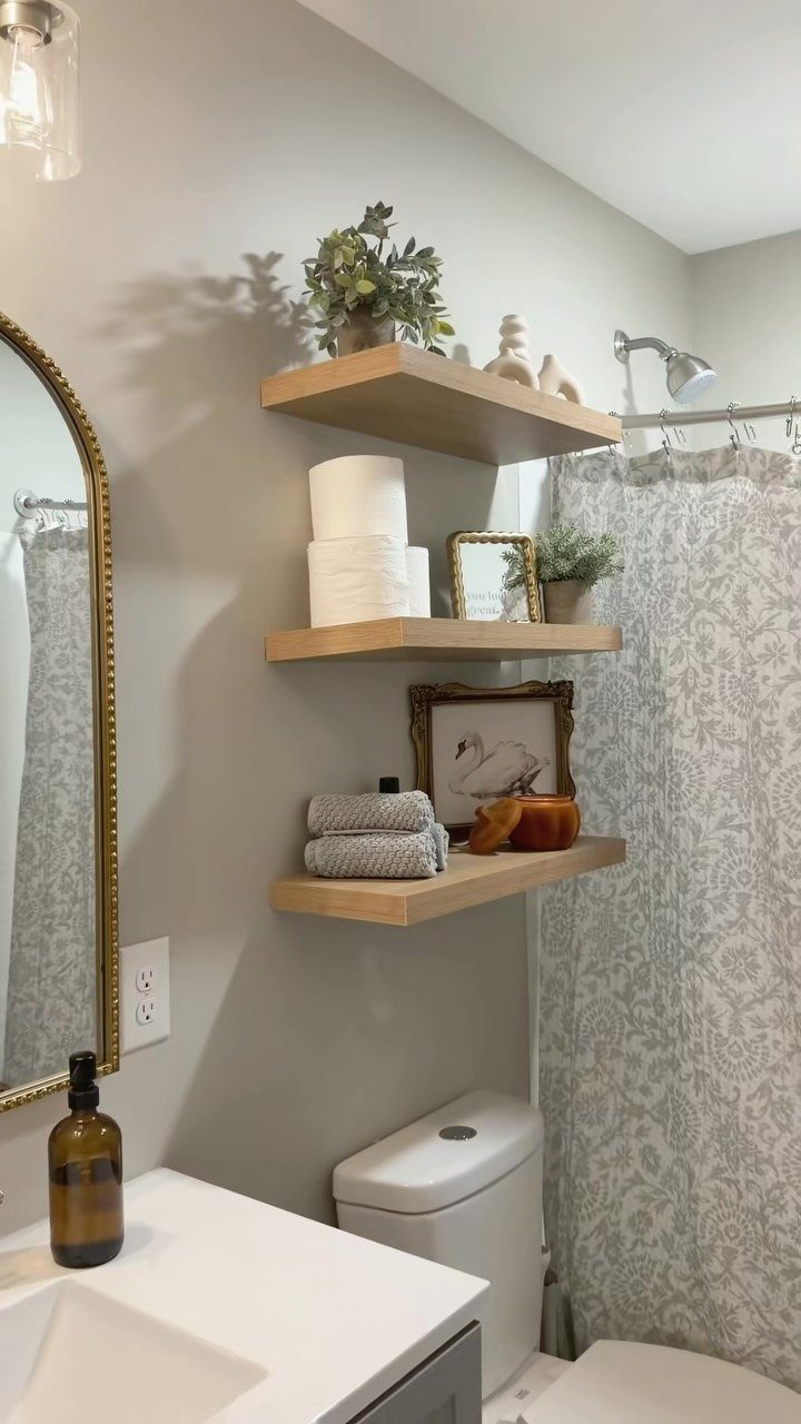 Three light-wood floating shelves with towels and decor above a white toilet.