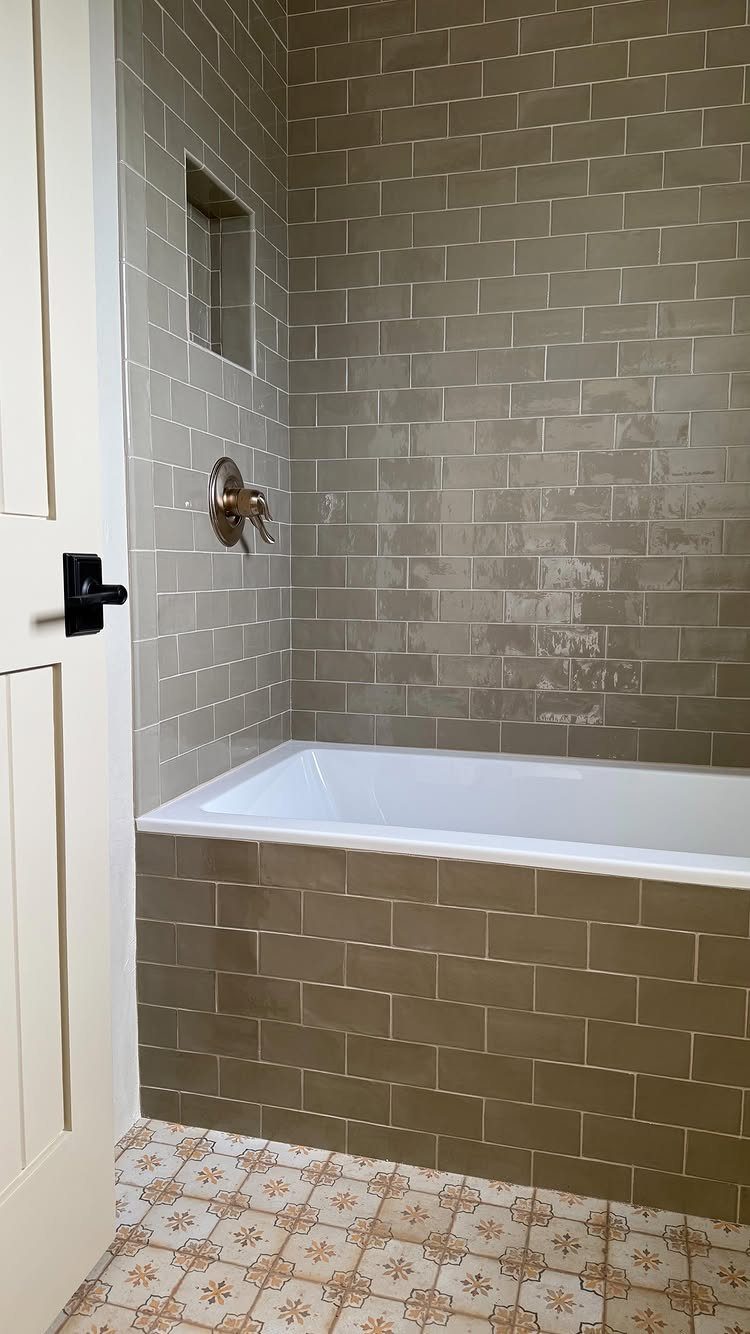 White alcove bathtub with grey subway tile walls and patterned floor tiles.