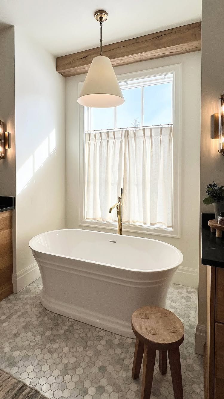 White freestanding bathtub in a bright bathroom with white café curtains on the window.