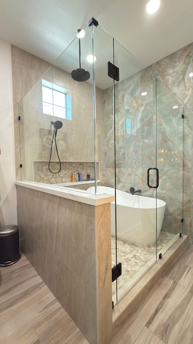 Frameless glass shower enclosure surrounding a white freestanding bathtub with marble tile walls.
