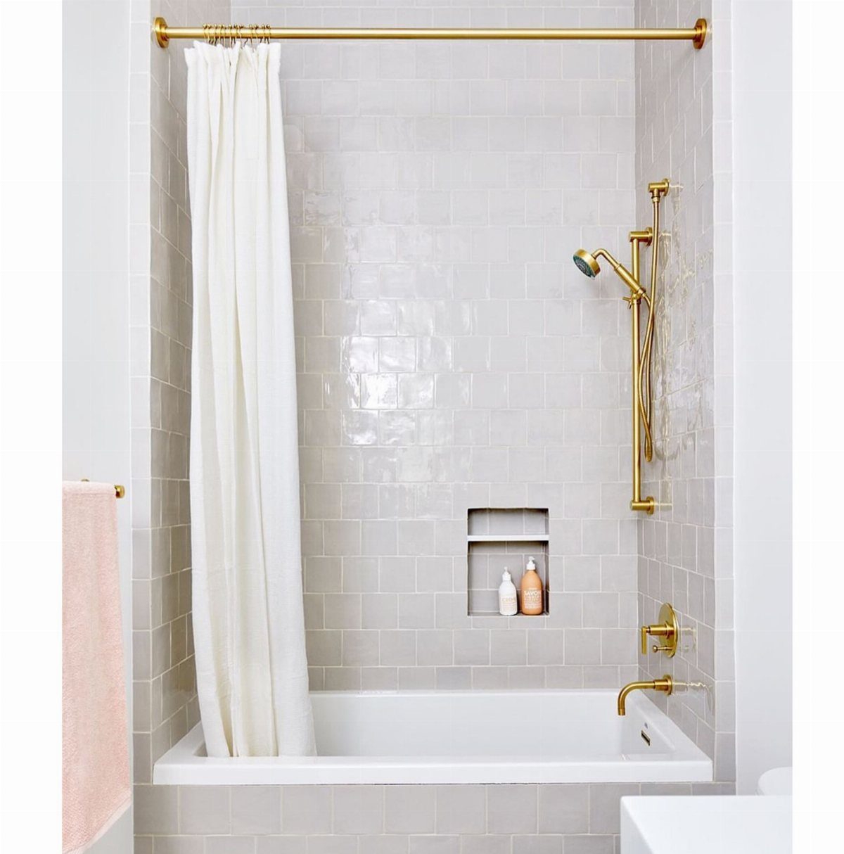 White bathtub with light grey tiled wall surrounds and gold shower fixtures.