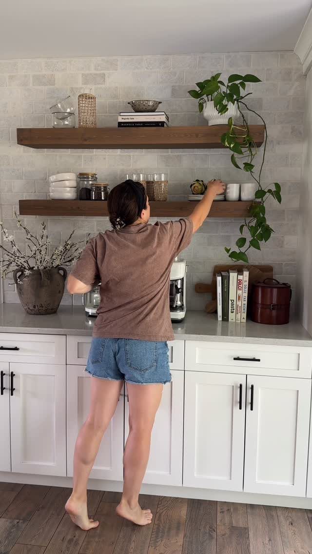 Woman reaching for a cup on wooden open shelving in a white kitchen.