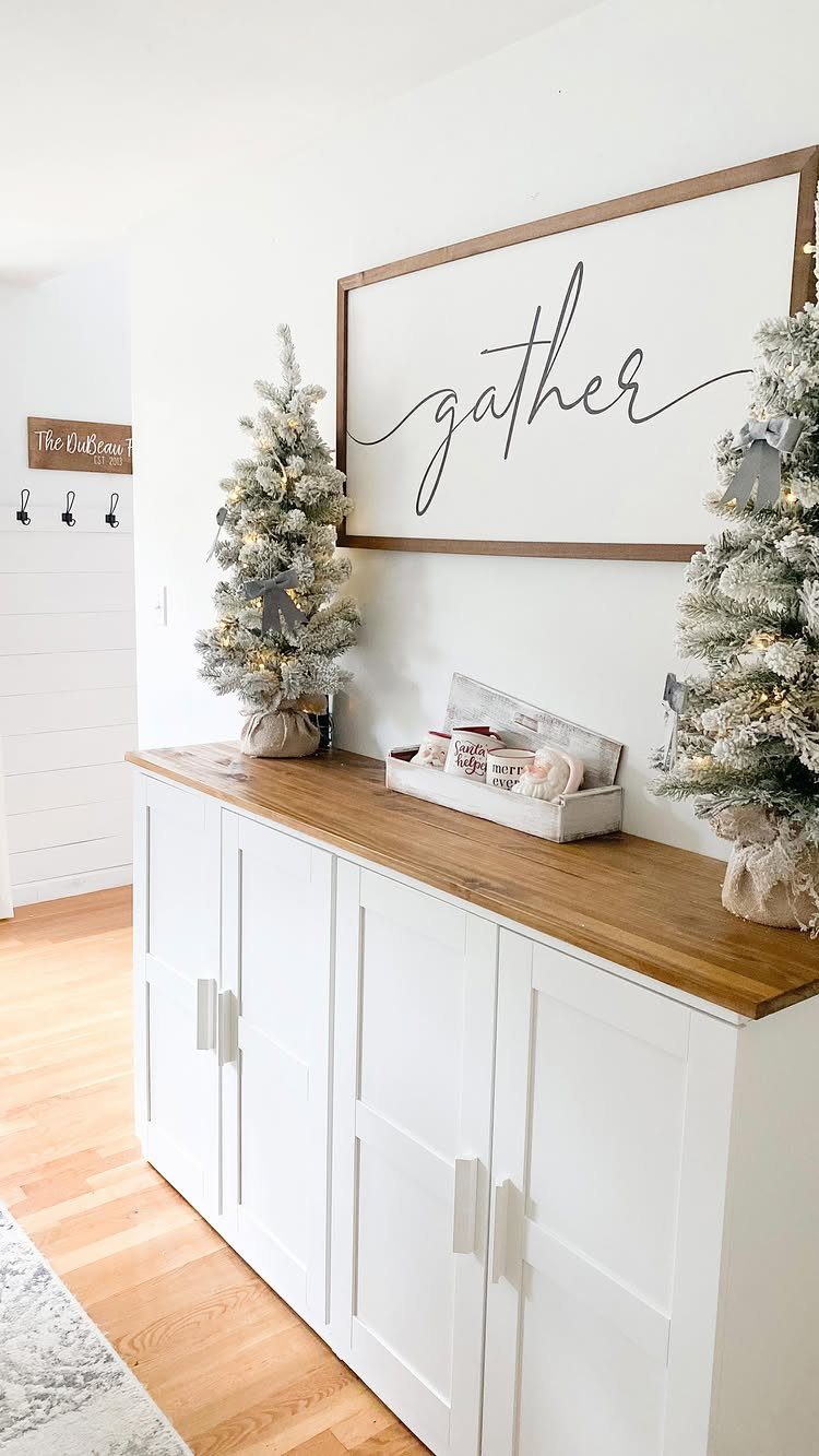 Two frosted mini evergreen trees on a white sideboard with a gather sign.