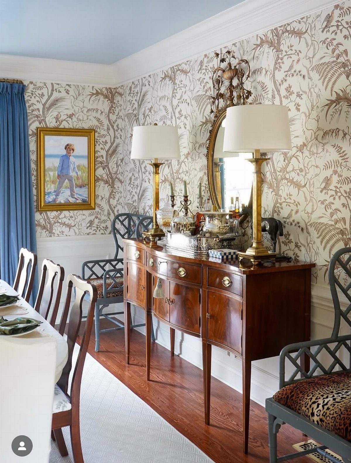 Wooden console table with gold lamps, a mirror, and artwork on floral wallpaper.