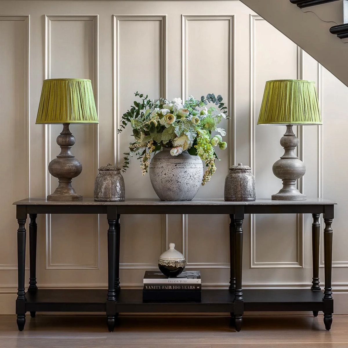 Slim dark wood console table with green lamps and floral arrangement against paneled wall