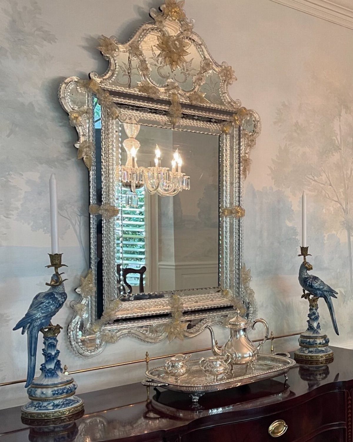 Ornate antique silver mirror hanging above a dark wood sideboard with peacock candle holders.