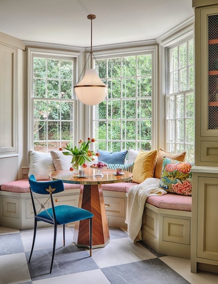 Dining nook with round wooden table and built-in bench inside a bright bay window.
