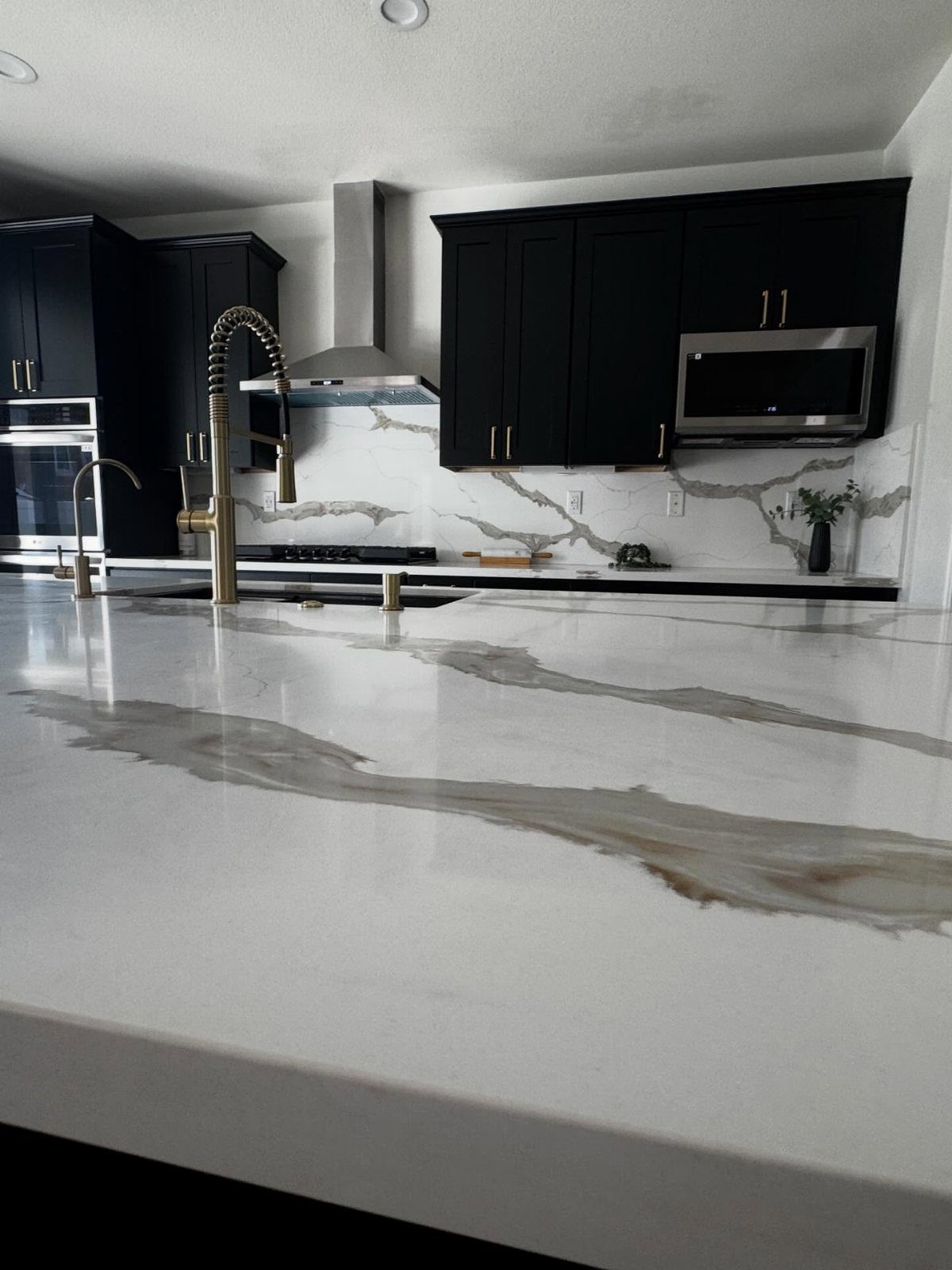 White marble kitchen countertops with grey veins and gold faucet against black cabinets.