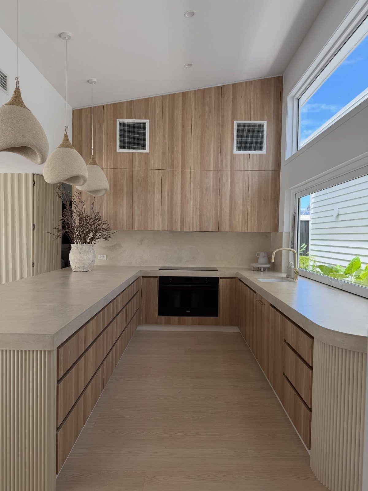 Light concrete countertops in a modern kitchen with light wood cabinets and woven lamps.
