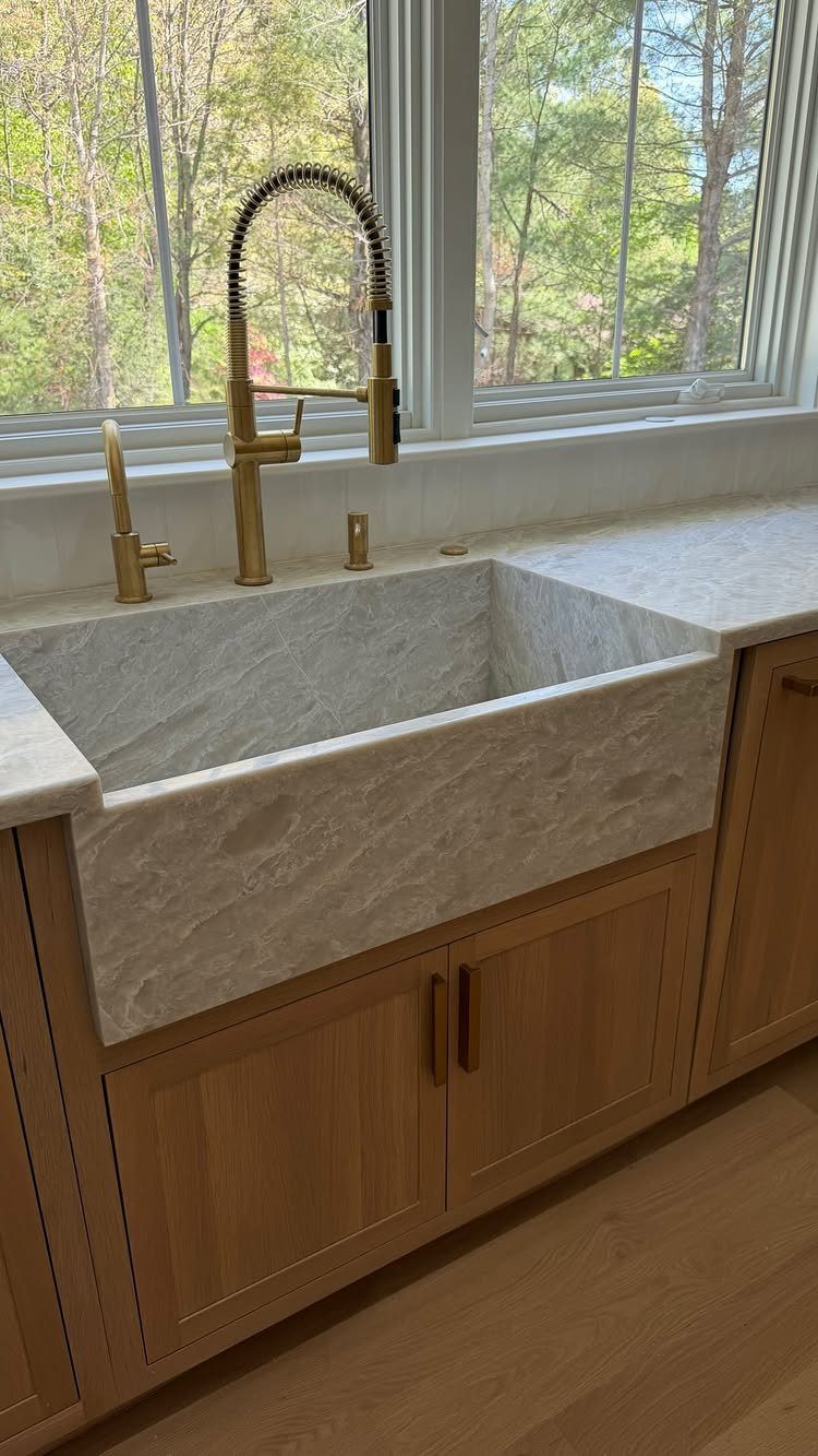 35 Farmhouse Kitchen Remodel Ideas Worth Stealing 2 White marble farmhouse sink with gold faucet and light wood cabinetry by a window.