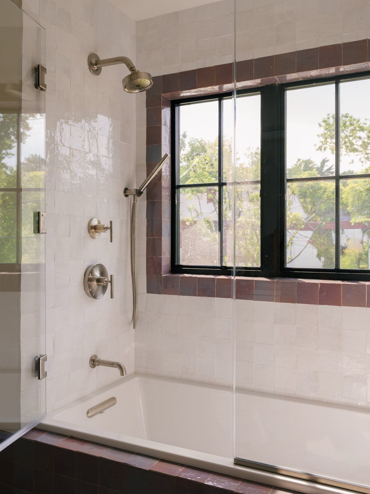 17 Jack and Jill Bathroom Remodel Ideas for Shared Spaces 4 White bathtub and shower with burgundy tile accents and a black-framed window.