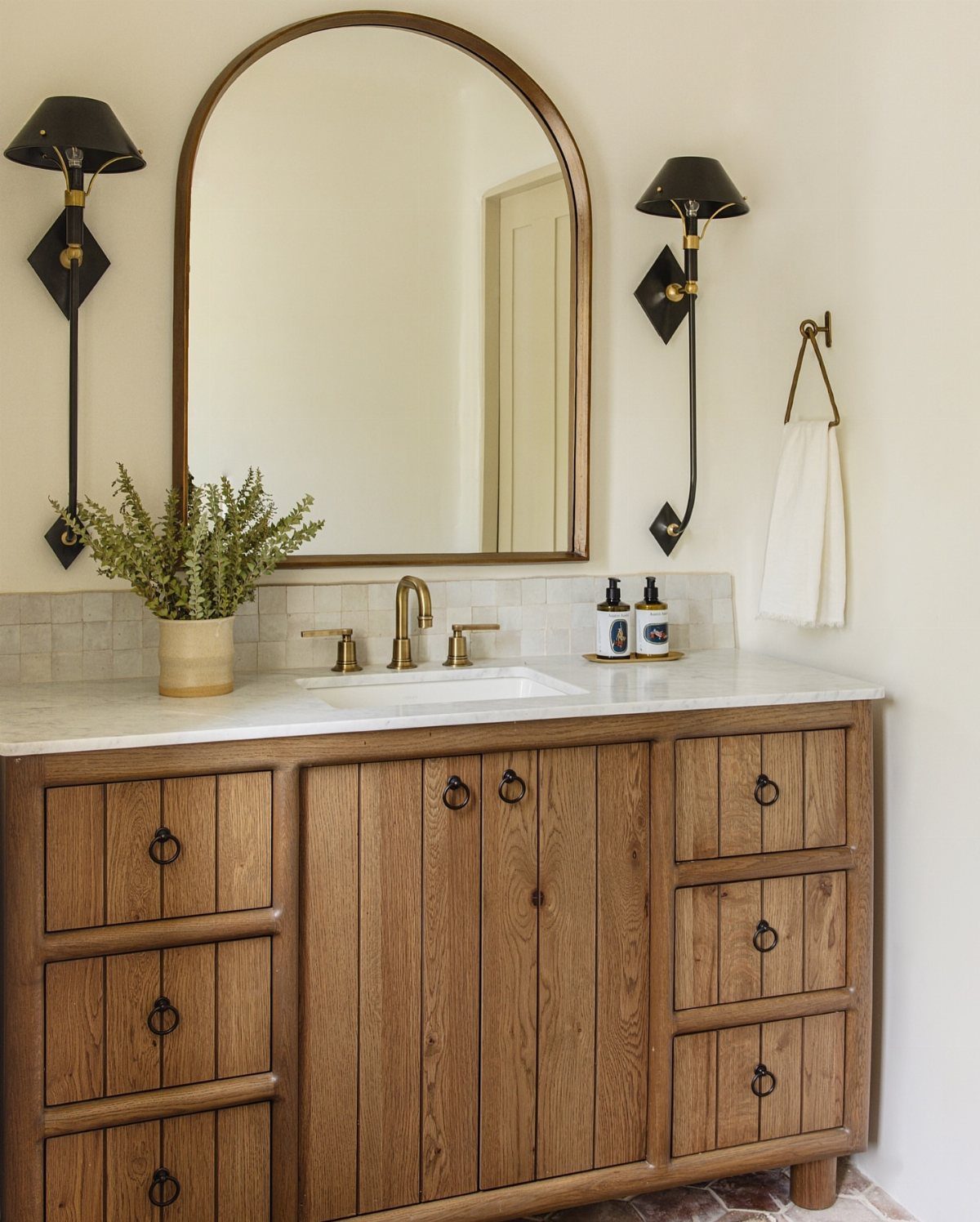 17 Jack and Jill Bathroom Remodel Ideas for Shared Spaces 11 Wooden bathroom vanity with white marble top, arched mirror, and black sconce lighting.