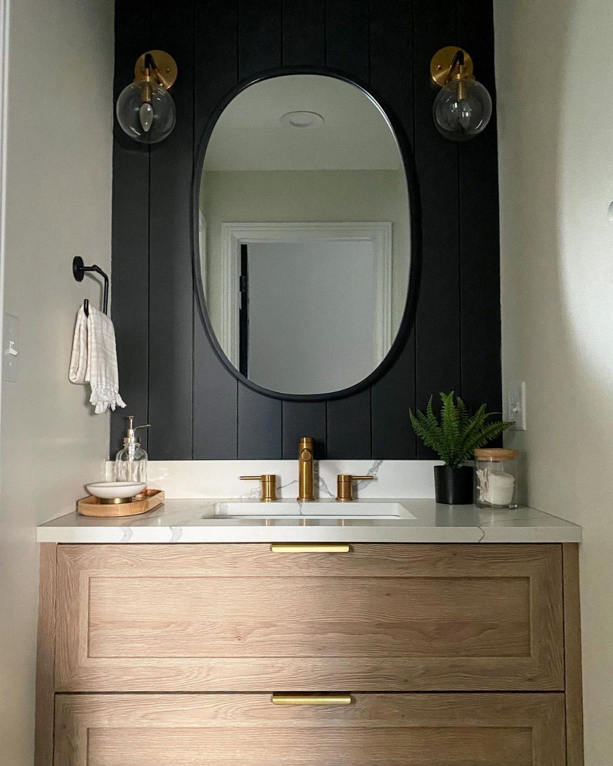 17 Jack and Jill Bathroom Remodel Ideas for Shared Spaces 6 Black shiplap accent wall with oval mirror and gold sconces above a wood vanity.
