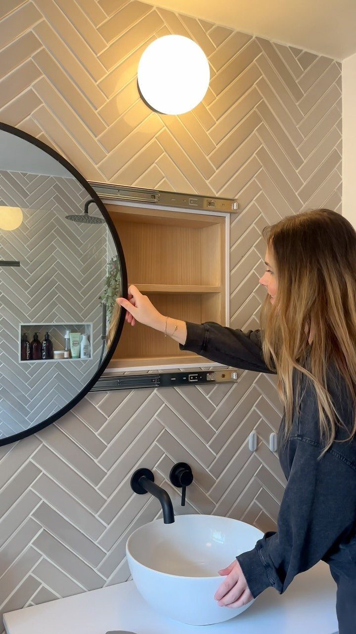 17 Jack and Jill Bathroom Remodel Ideas for Shared Spaces 3 Woman opening a circular mirror to reveal recessed wooden shelving in a tiled bathroom.