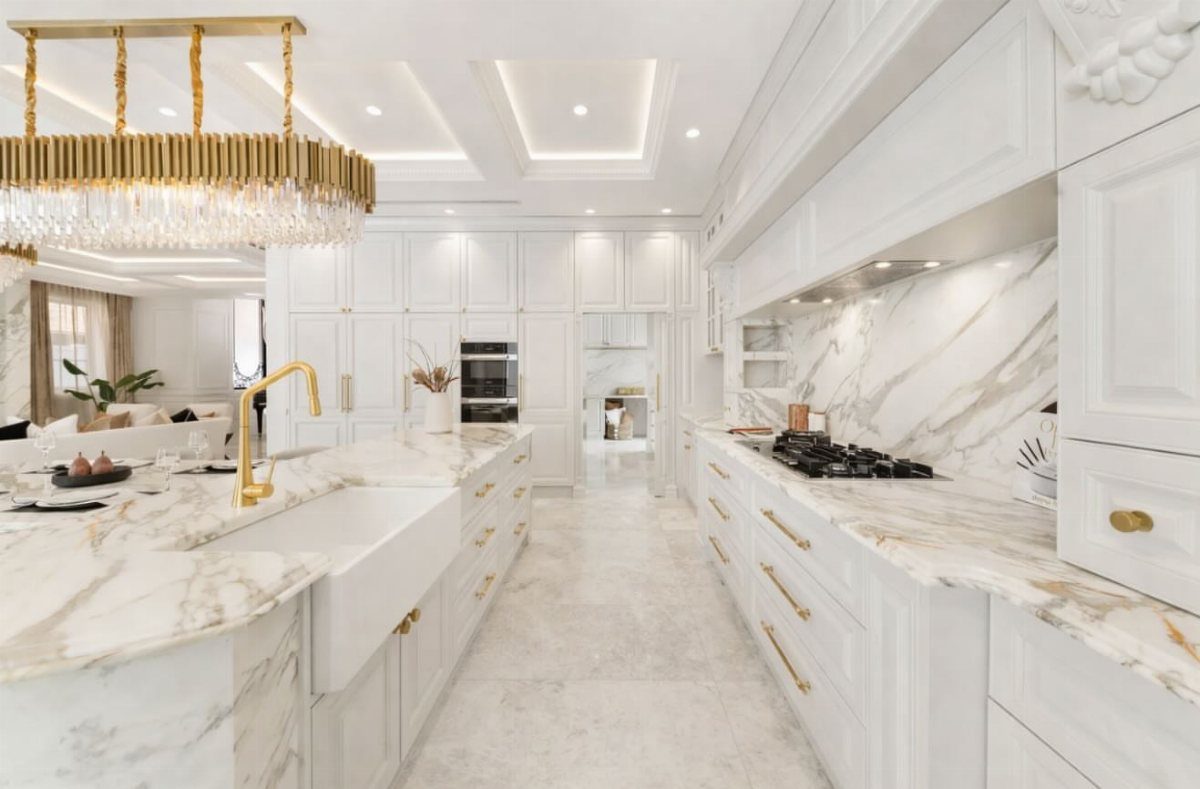 White ornate kitchen with marble countertops, gold fixtures, and a large crystal chandelier.