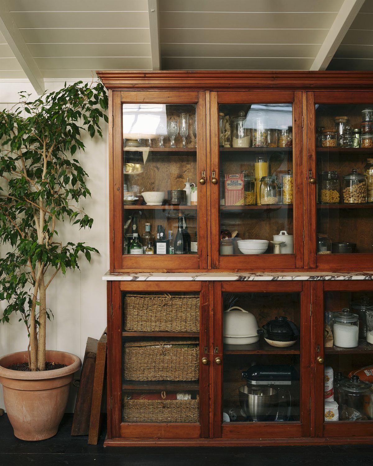Vintage wooden hutch with glass doors and woven baskets next to a potted plant.