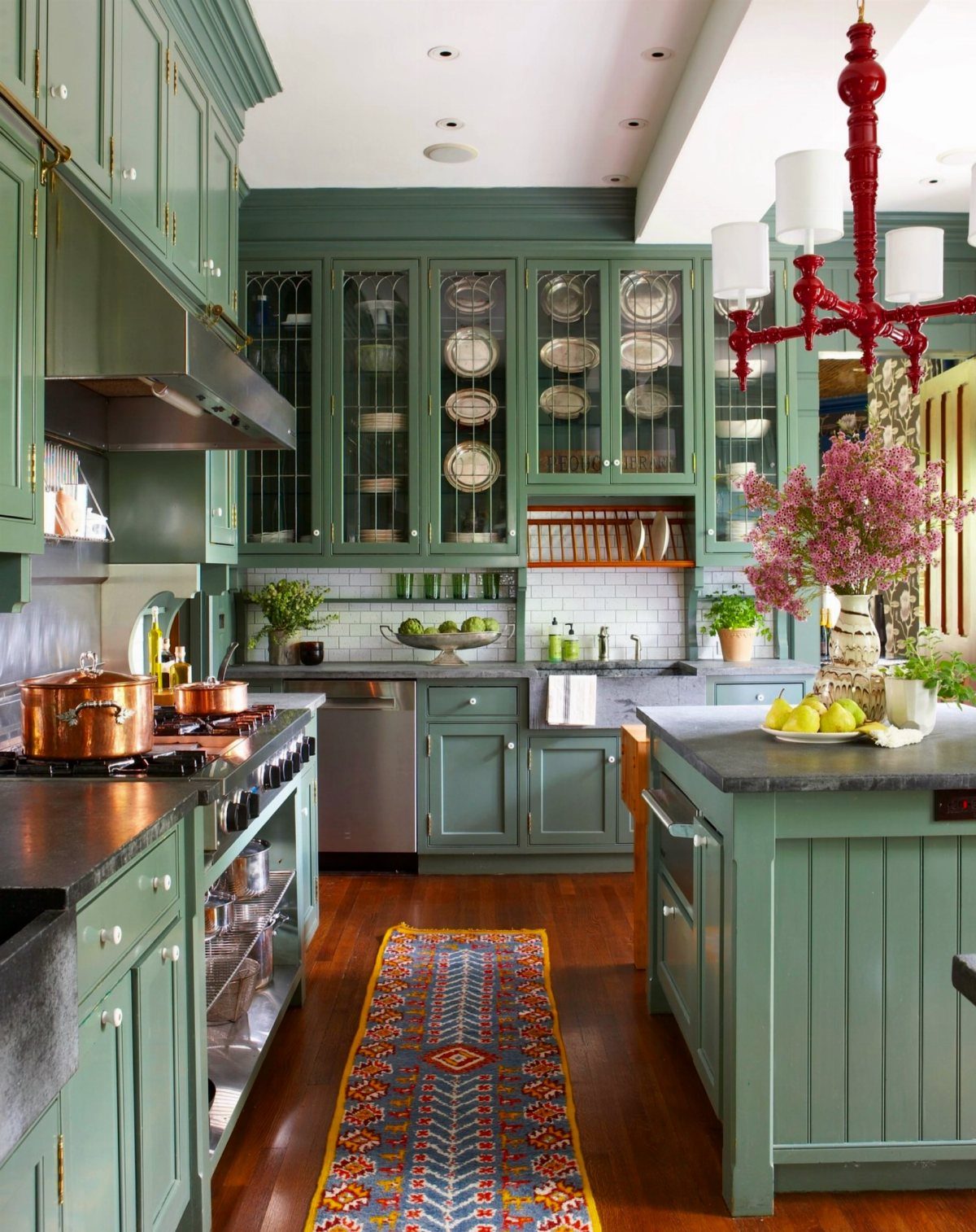 Sage green kitchen cabinets with a contrasting red chandelier and colorful runner rug.