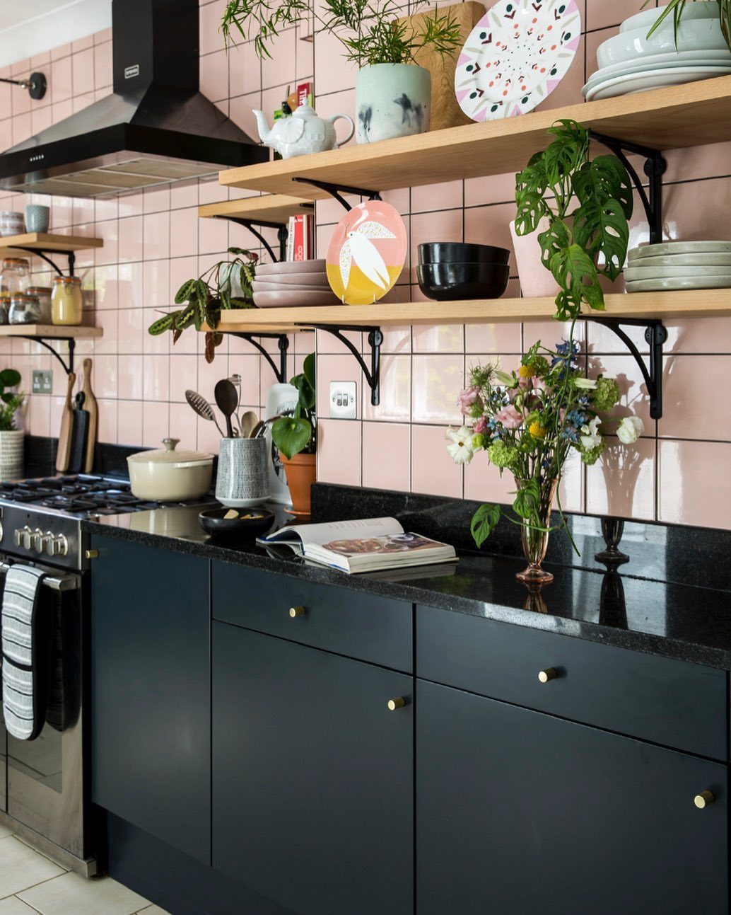 Black kitchen cabinets and countertops against a pink tiled wall with wooden shelves.