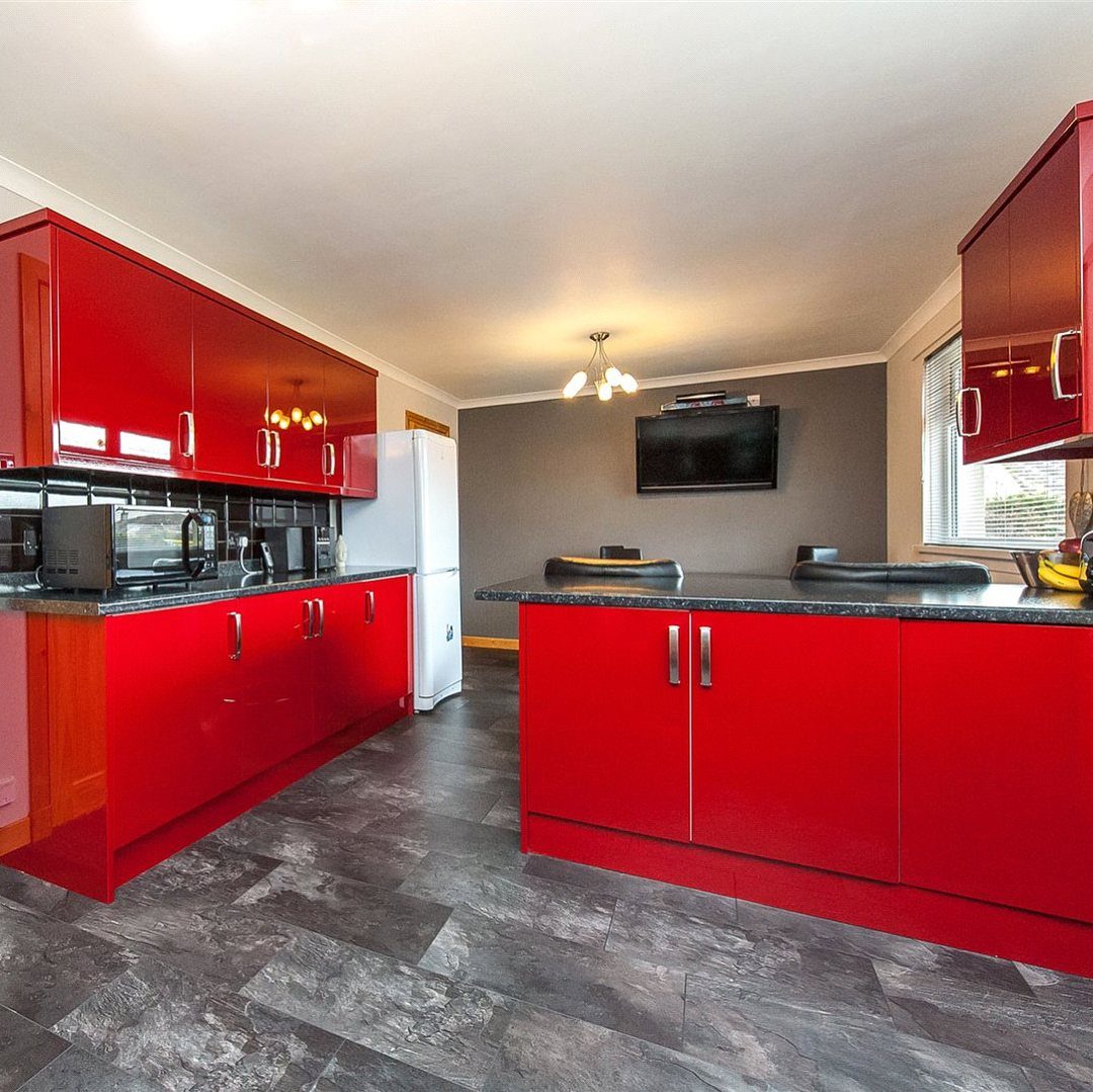 Glossy red kitchen cabinets and island with black countertops and grey tiled flooring.