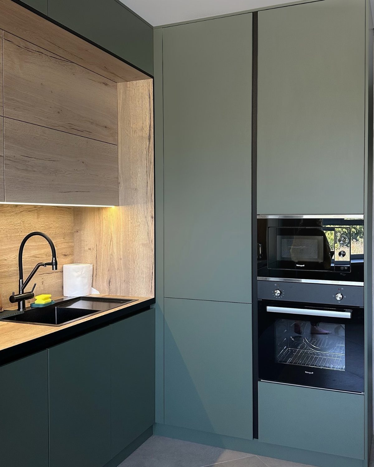 Sage green kitchen cabinets with light wood accents and black appliances.