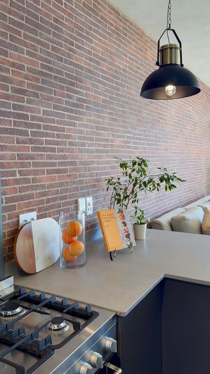 Red brick wall in a kitchen with grey countertops and black pendant lighting.