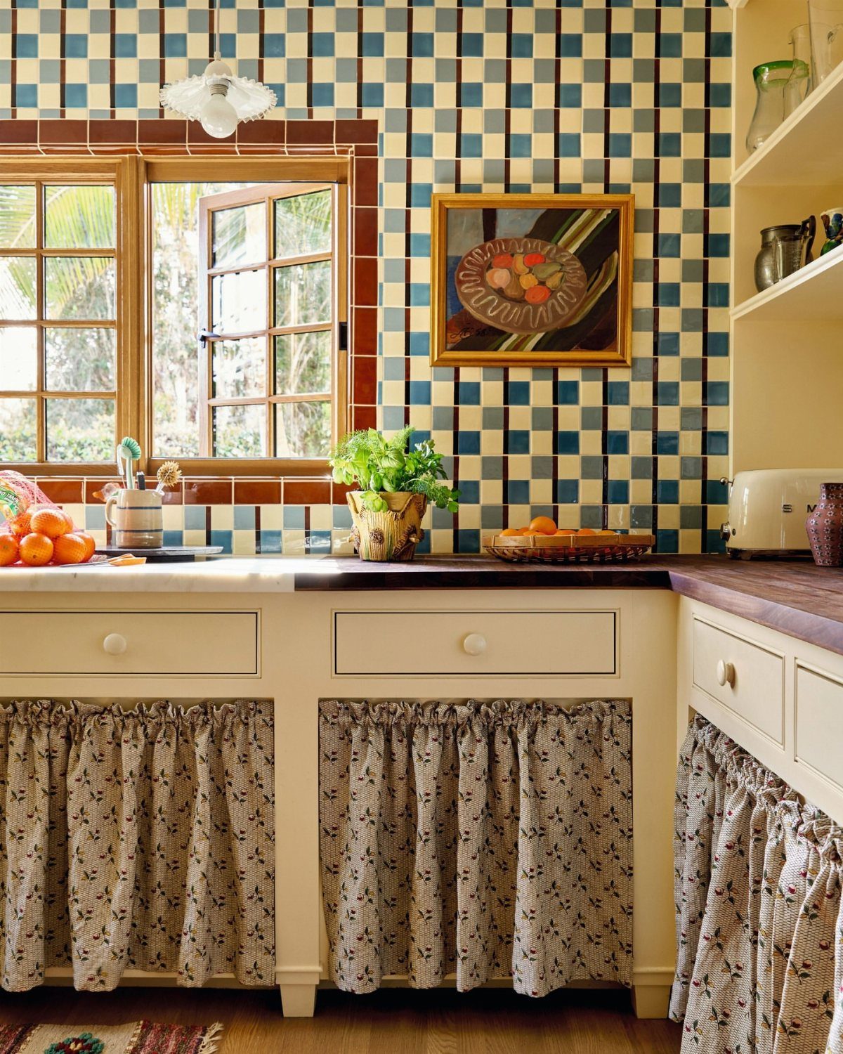 Cream kitchen cabinets with floral fabric skirts under a wooden countertop and checkered tiles.