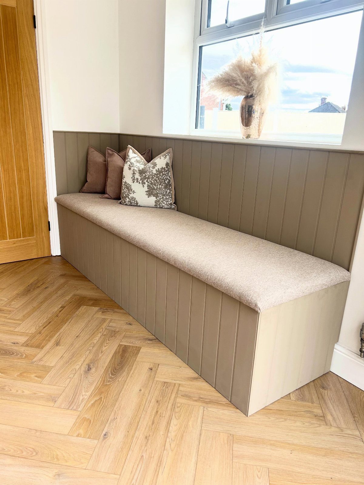 Built-in kitchen nook bench with grey paneling and beige upholstery on herringbone floors.
