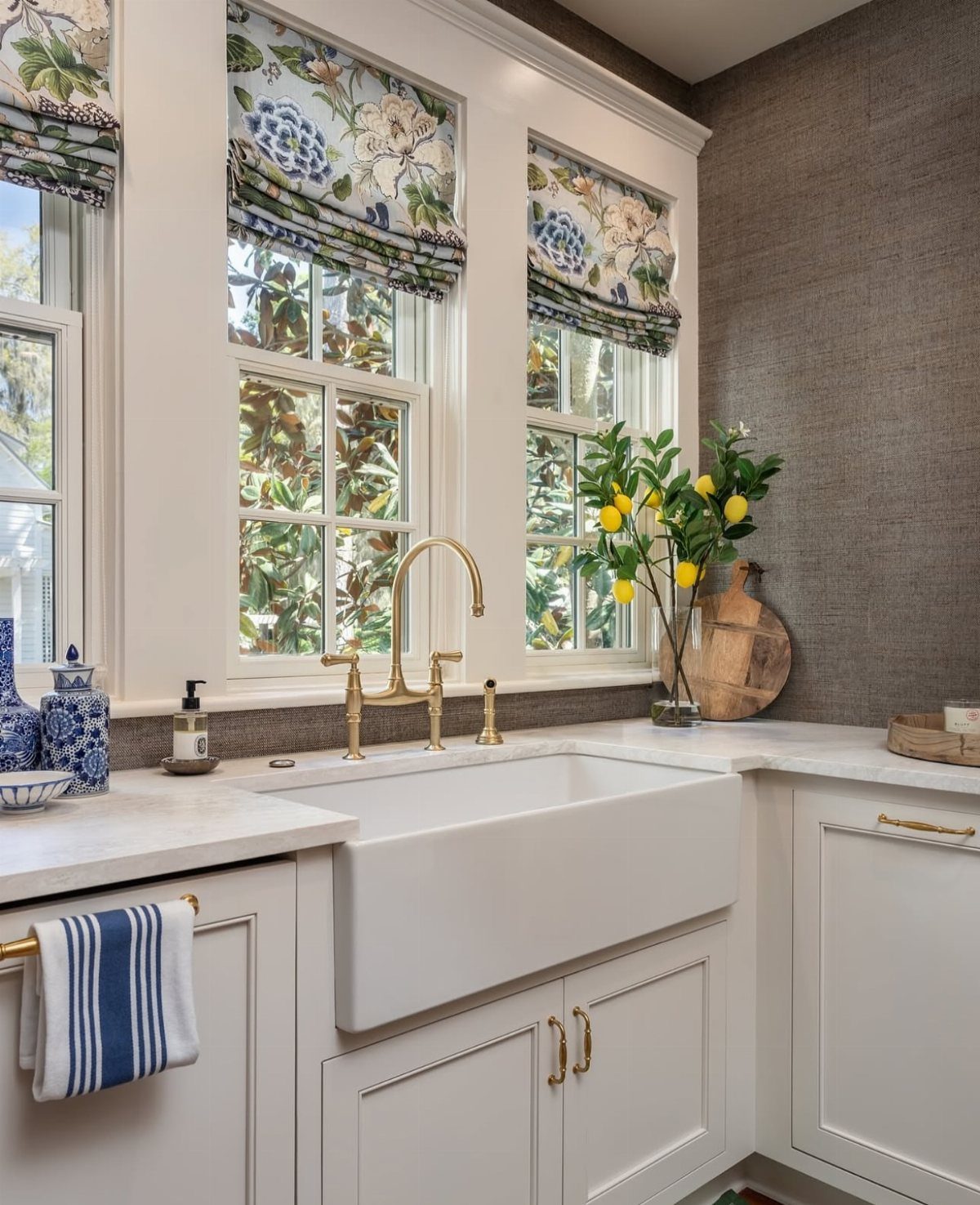 12 Kitchen Updates on a Budget, From Hardware to Pantry Jars 11 Floral Roman shades on kitchen windows above a white farmhouse sink and gold faucet.