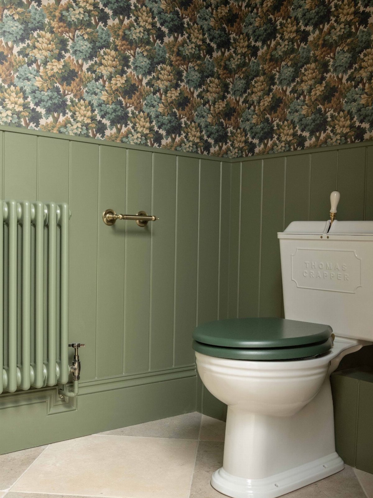 White toilet with green lid against sage green wainscoting and forest wallpaper.