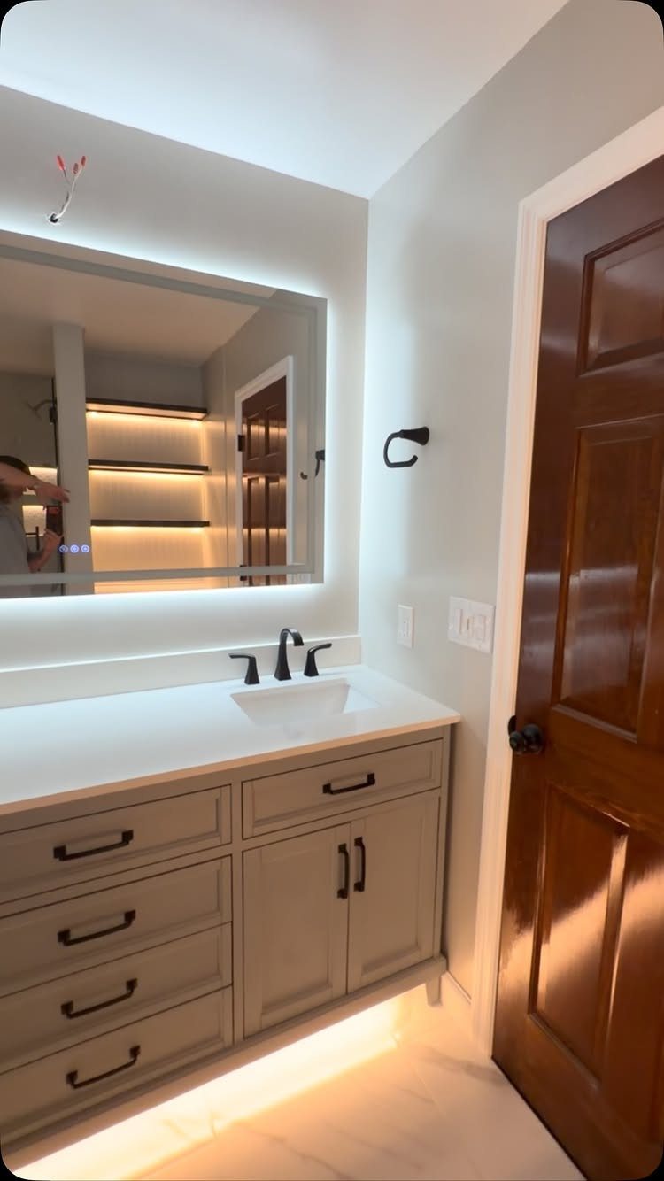 Grey bathroom vanity with white countertop, undermount sink, and a large backlit mirror.