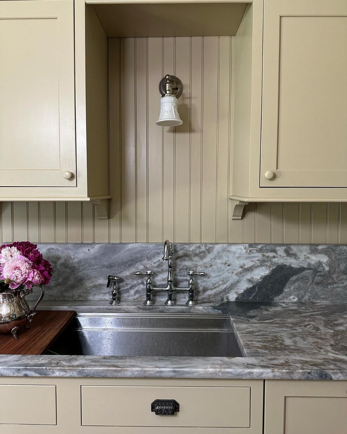 Stainless steel kitchen sink with marbled countertops and cream-colored cabinetry and wall paneling.