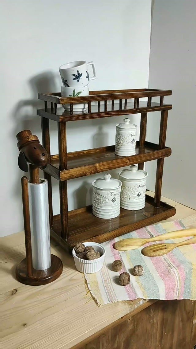 Three-tier wooden kitchen shelf holding white ceramic jars and mugs on a countertop.