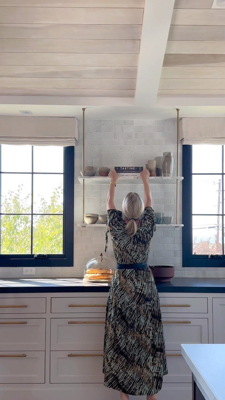 Woman placing a book on white open kitchen shelves against a tiled wall.