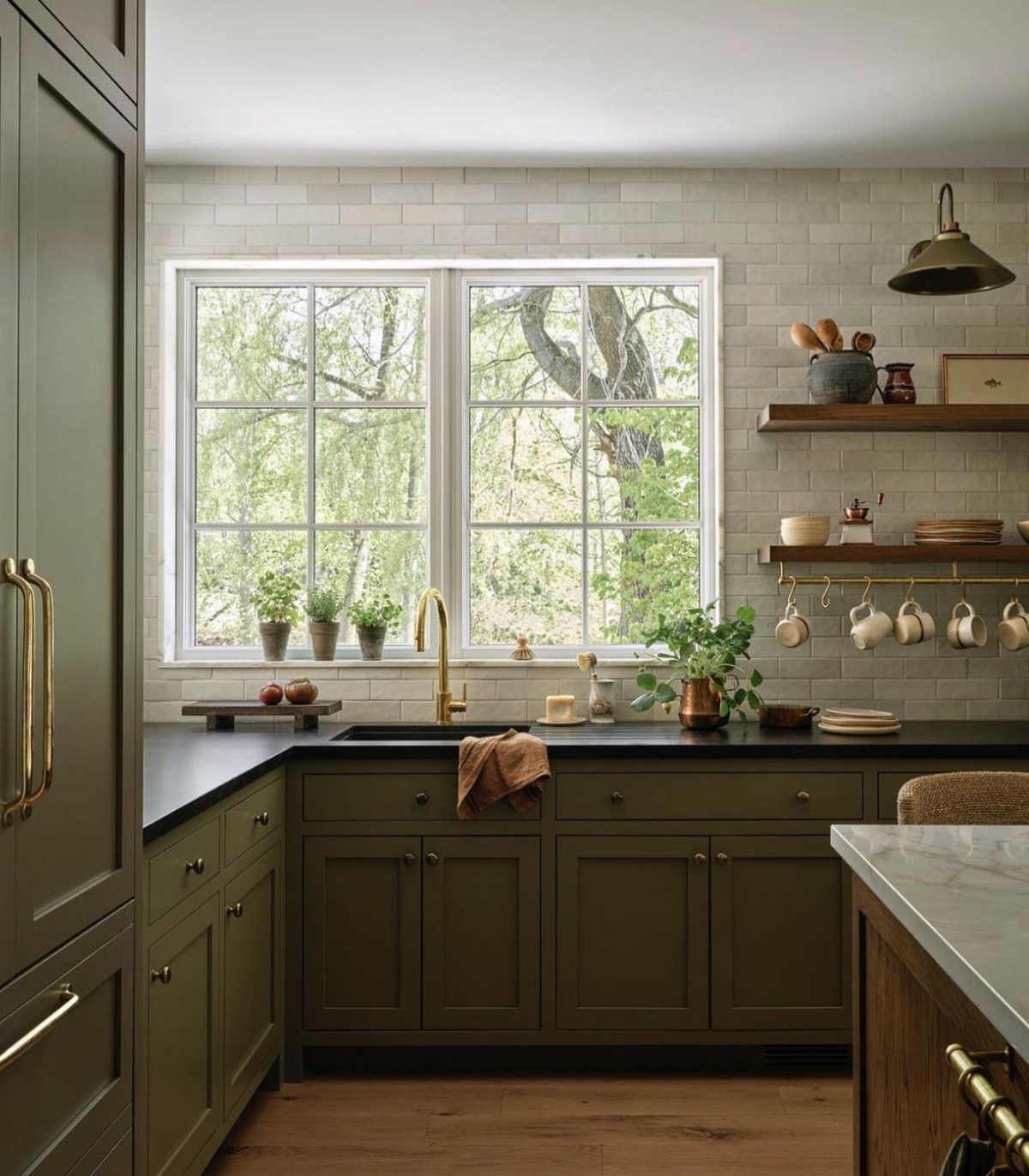 26 Sage Green Kitchen Ideas Using Wood, Brass, and Marble 20 Sage green kitchen cabinets with black countertops and gold hardware near a window
