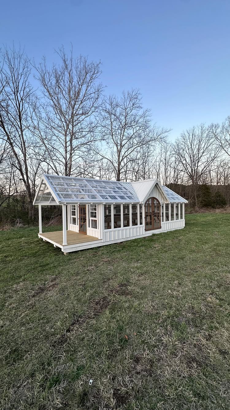 7 Shed with Porch Ideas for Storage, Studios and More 6 White garden shed with a glass roof and a small wooden porch in a field.